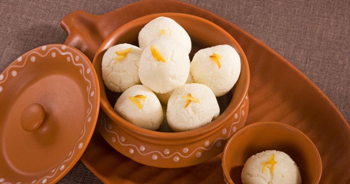 Clay pot filled with white rasgulla sweets, each topped with saffron strands, on a terracotta serving tray.