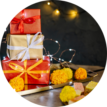 Stack of colorfully wrapped gift boxes with red and gold ribbons, glowing fairy lights, and yellow marigold flowers on a festive table.