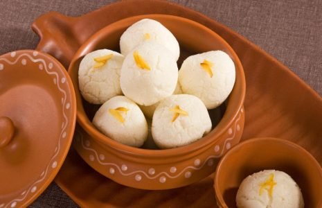 Clay pot filled with white rasgulla sweets, each topped with saffron strands, on a terracotta serving tray.